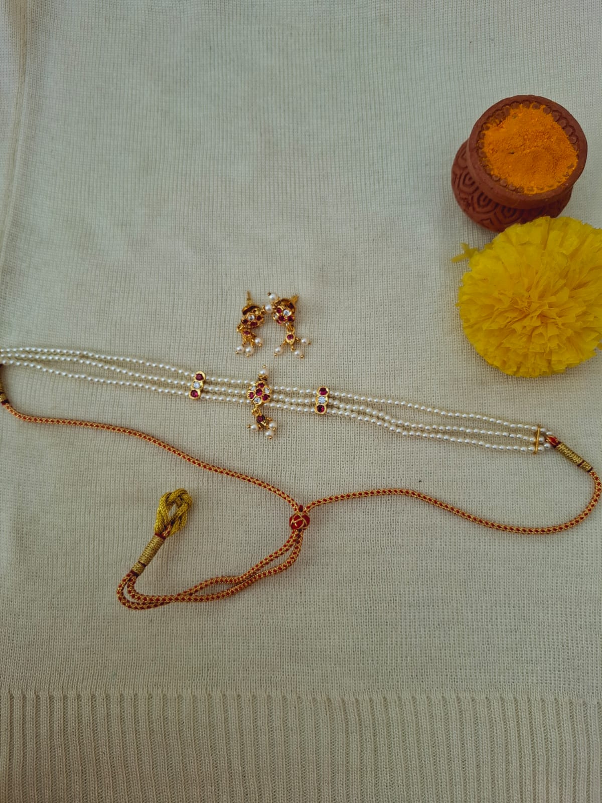 Small flower-shaped traditional Indian jewelry pieces in copper and yellow tones, including a necklace, earrings, and a pompon accent, arranged on a light gray knitted fabric.