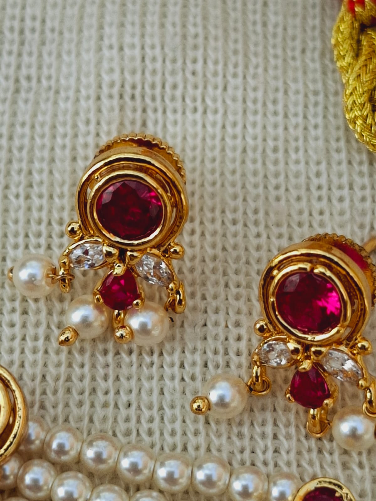 Ornate gold-toned earrings with vibrant red gemstones and shimmering white pearls, set against a textured fabric background.