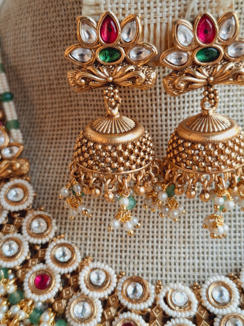 Ornate golden necklace with vibrant red and green gemstones, set against a woven textile background in the Mugdha Jewellery Store.