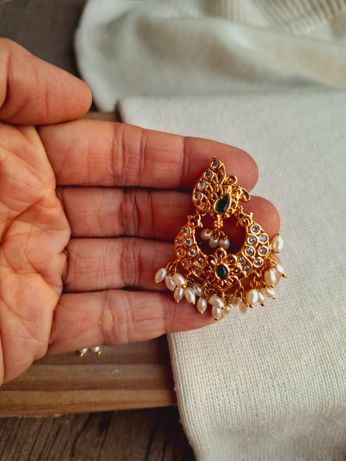 Ornate gold earrings with pearl tassels and a floral design, displayed in a hand against a neutral background.
