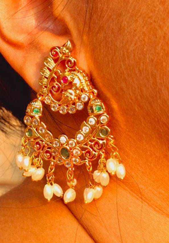 Ornate peacock earrings adorned with vibrant gems and dangling pearls, showcased on a striking red hair background in the Mugdha Jewellery Store.