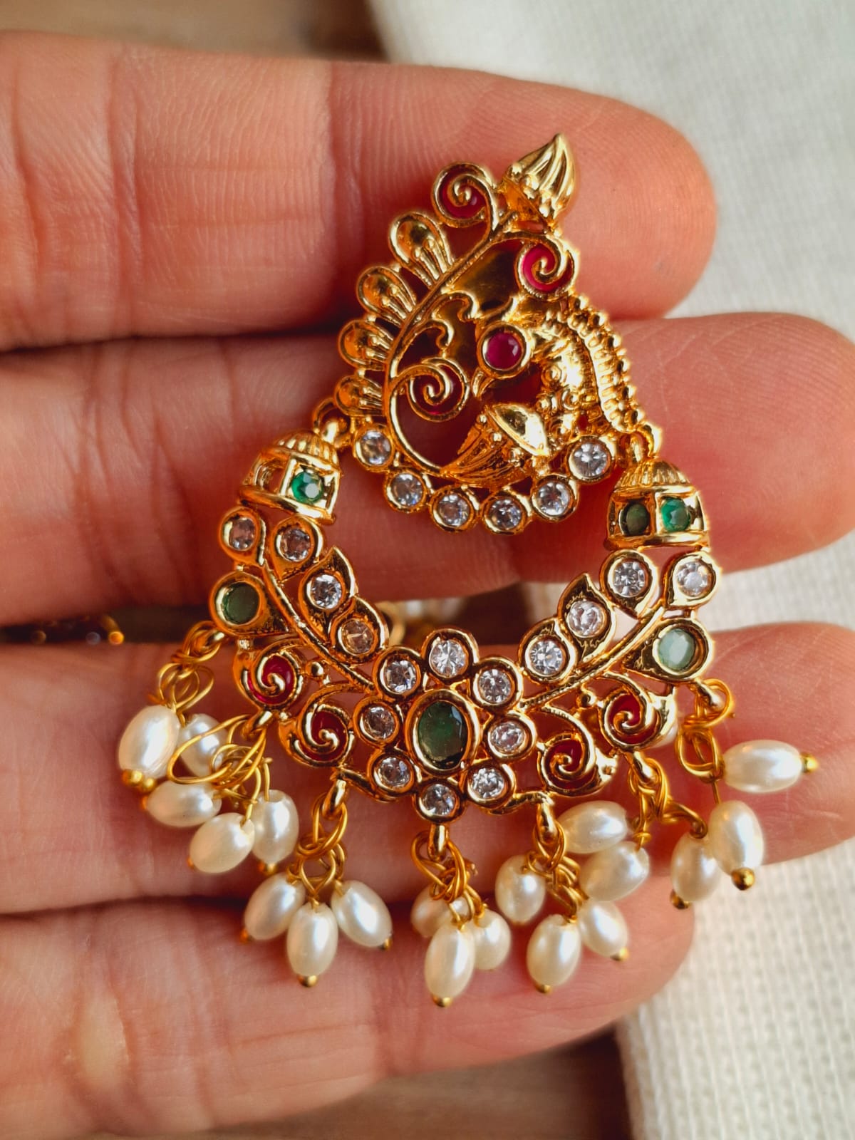 Elegant gold-toned peacock-shaped earrings adorned with sparkling rhinestones and dangling pearl-like beads, showcased in the palm of a hand.