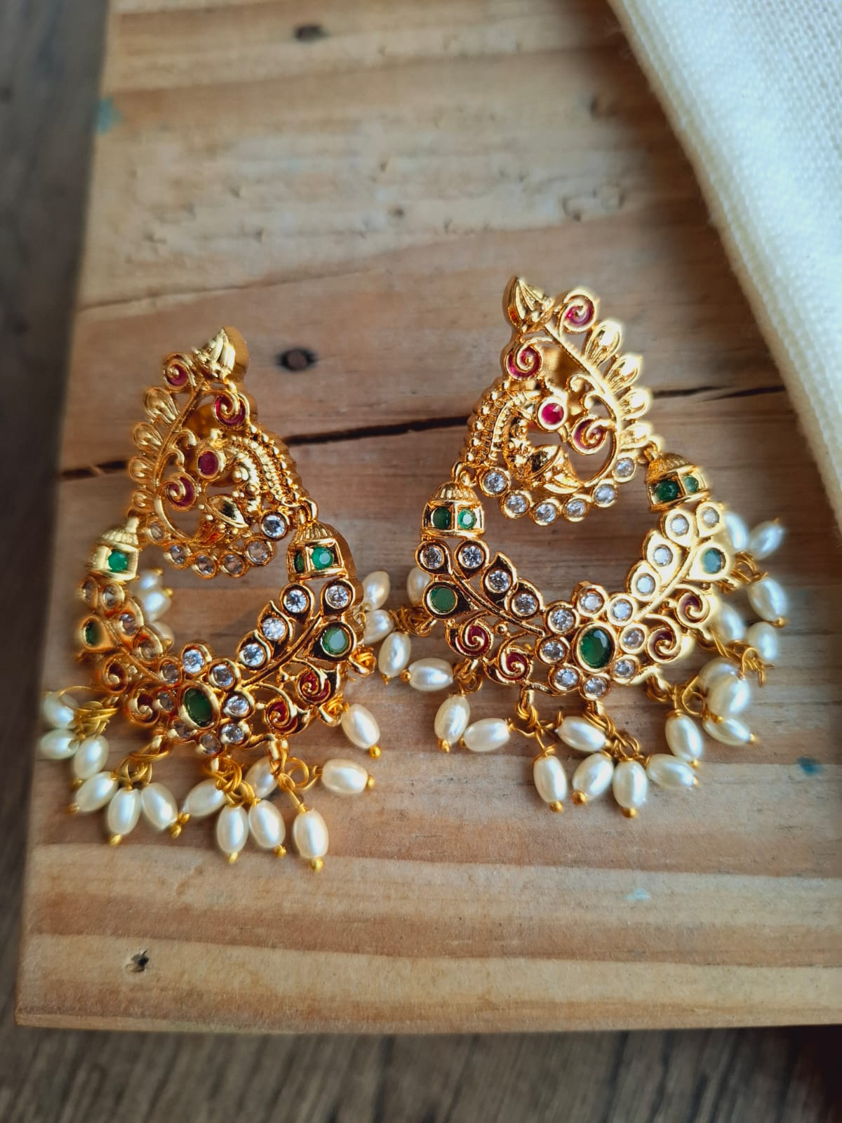 Ornate gold-toned peacock-shaped earrings adorned with vibrant multicolored gems and pearls, displayed on a rustic wooden surface.