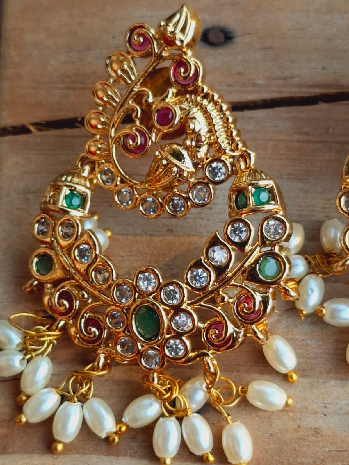 Ornate gold-toned peacock-shaped earrings accented with colorful gemstones and pearls, displayed against a wooden background.