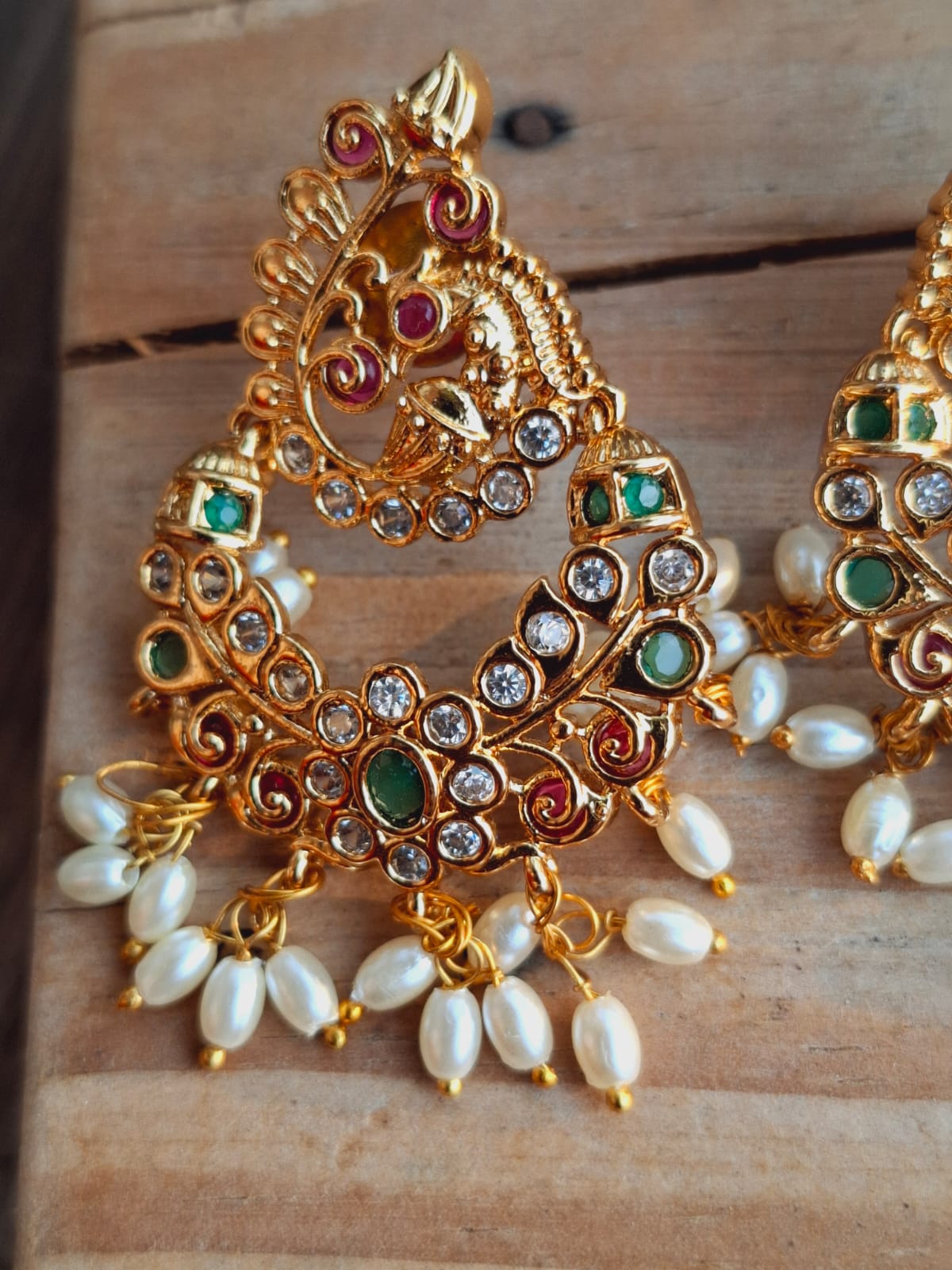 Ornate gold-toned peacock earrings with intricate filigree design, studded with colorful gemstones and dangling white pearls, displayed on a wooden background.