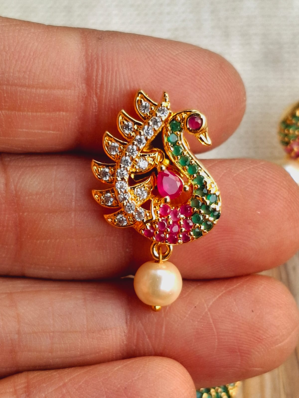 Ornate peacock pendant in gold tone with sparkling jewels and a pearl accent, held in a person's hand.