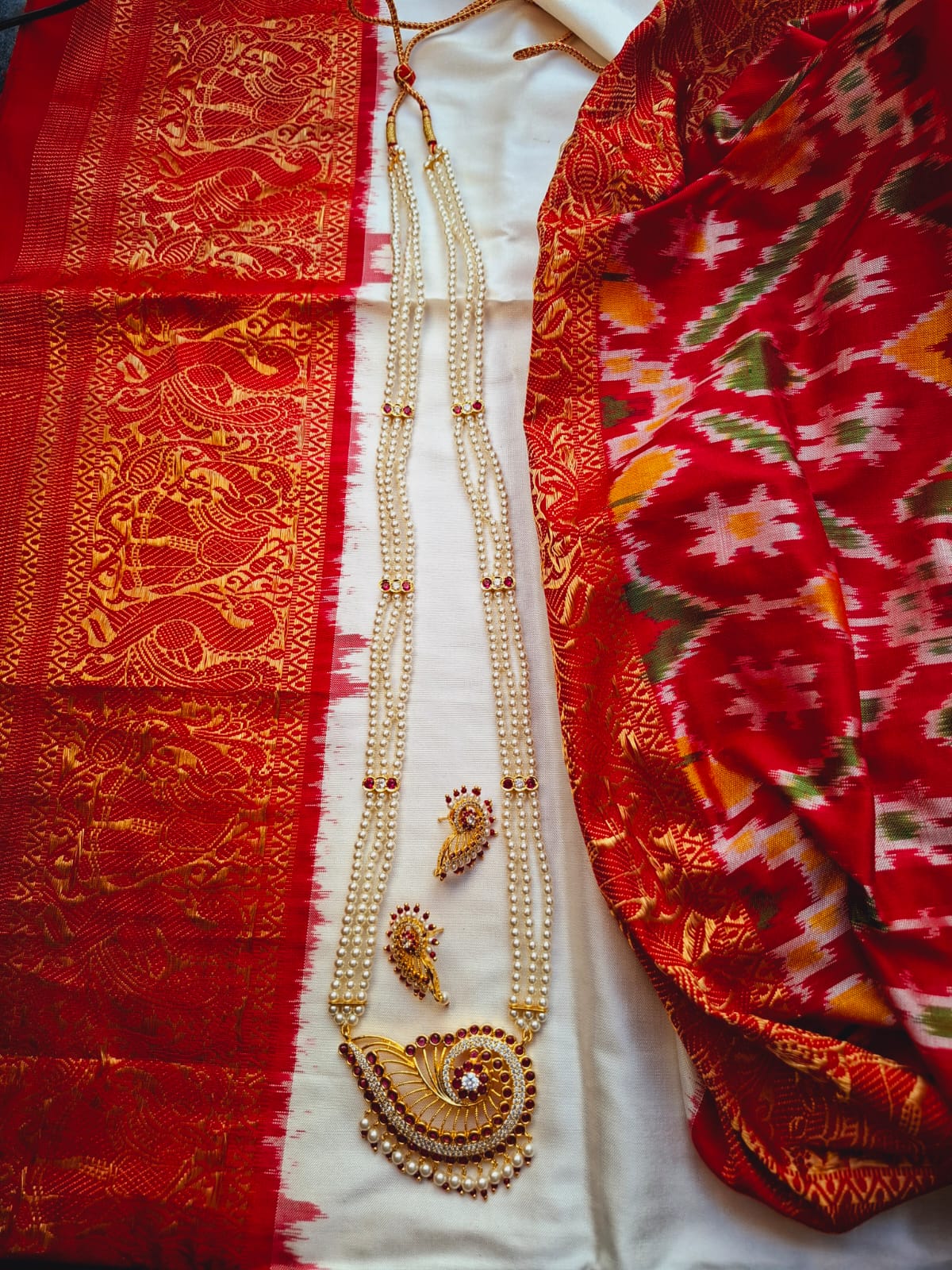 Elegant Maharani designer long necklace set on display, featuring intricate golden embroidery on a vibrant red and orange fabric backdrop, with gold-toned jewelry pieces showcased on a white surface.