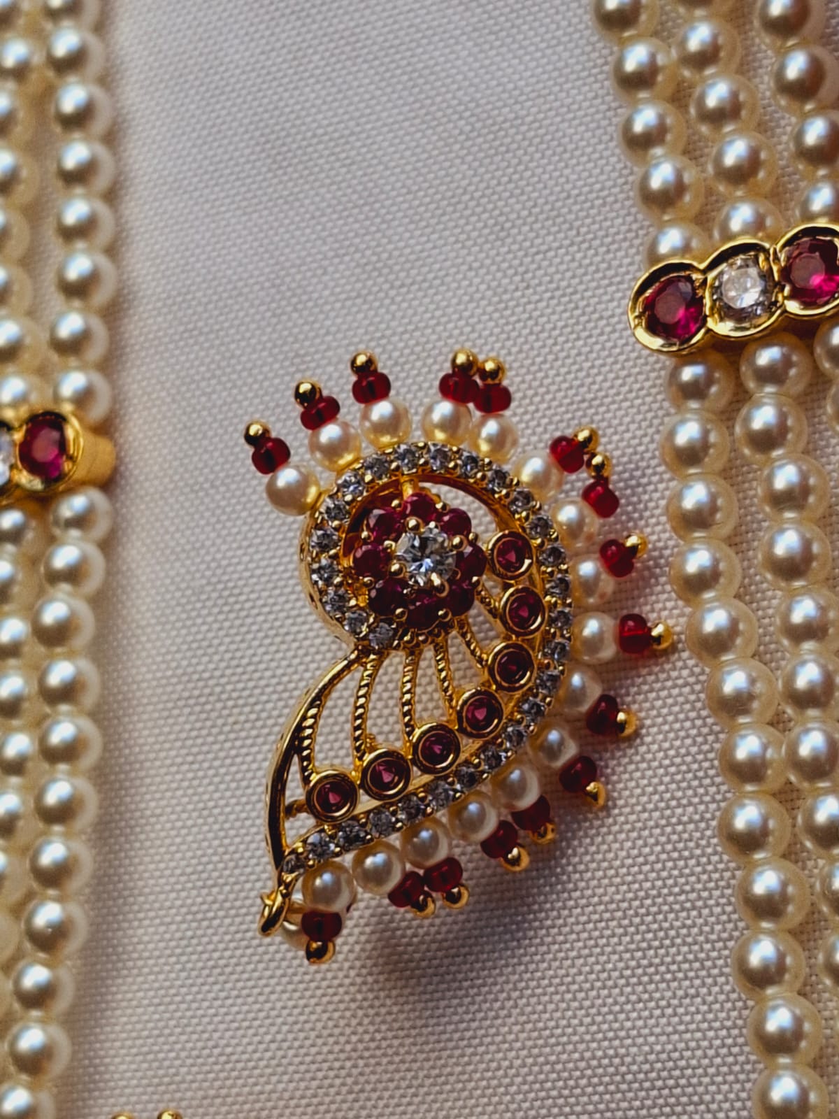 Stunning Maharani Designer Long Haar: Ornate golden brooch with intricate ruby and pearl details, set against a backdrop of lustrous pearl necklaces in the Mugdha Jewellery Store collection.
