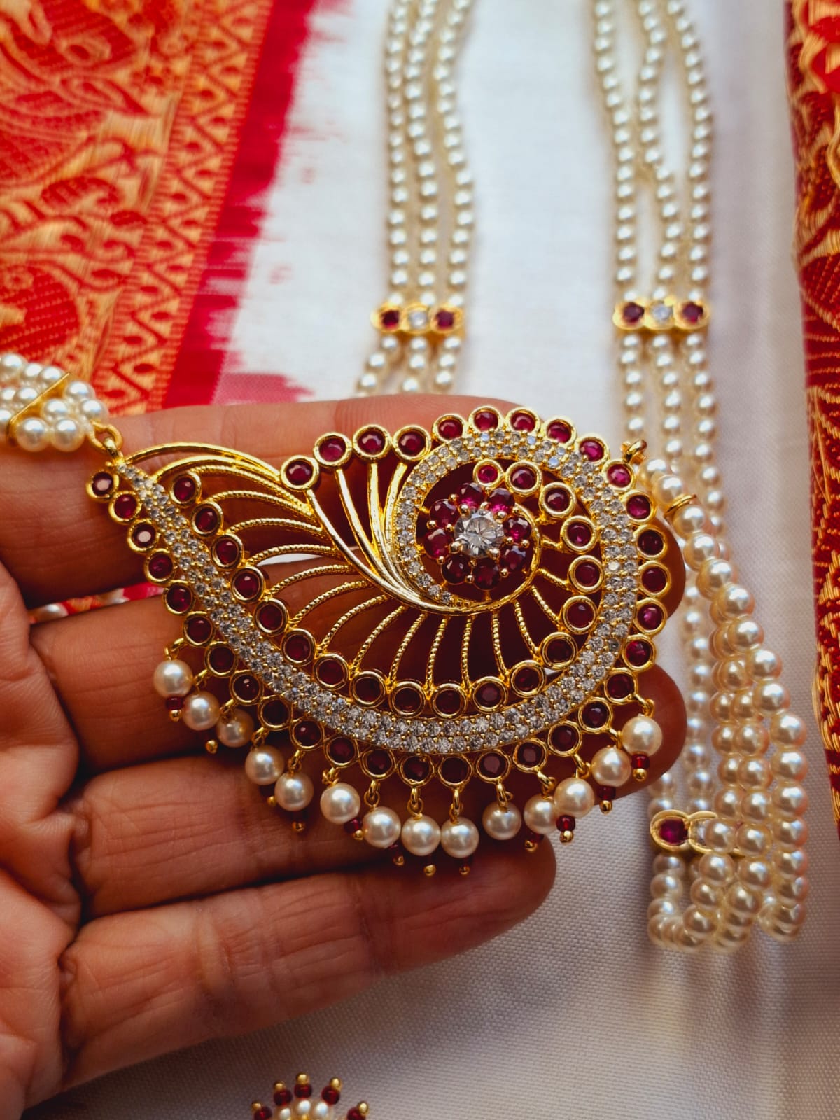 Intricate gold-toned necklace featuring a bold, jewel-encrusted peacock design accented with lustrous pearls, set against a vibrant red and gold patterned fabric backdrop.