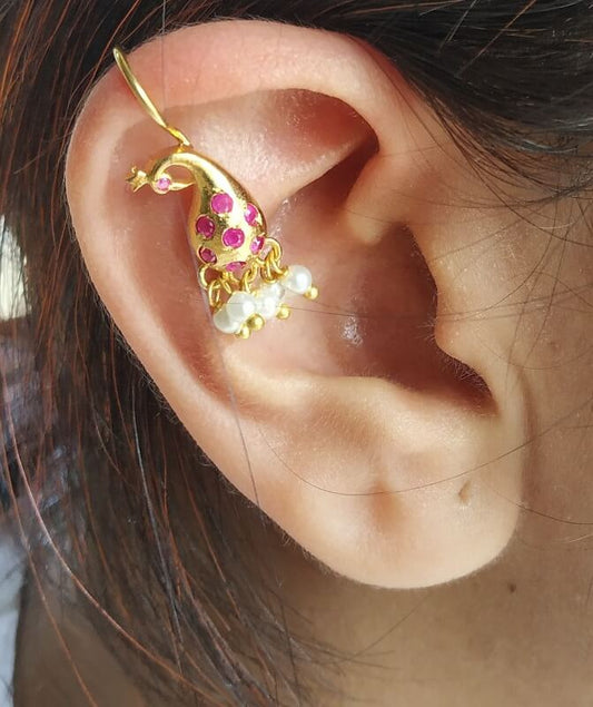 Elegant gold and ruby peacock-shaped ear cuff with hanging pearls, displayed on a human ear against a dark background.