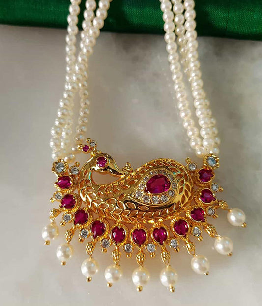 Elegant gold-toned peacock-shaped pendant with vibrant red gemstones and lustrous pearl accents, displayed on a multi-strand pearl necklace on a green velvet background.