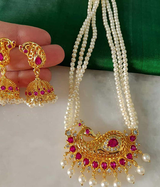 Elegant pearl and ruby necklace with matching jhumka earrings, displayed on a green fabric background. The necklace features a stunning golden peacock-shaped pendant encrusted with vibrant ruby stones, surrounded by multiple strands of lustrous pearls. The earrings complement the necklace with their ornate golden design and ruby embellishments.