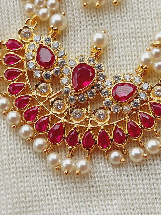 Elegant gold-toned designer necklace with intricate ruby-red and crystal gemstone accents, adorned with lustrous pearl beads, showcased against a soft, neutral knitted fabric background.