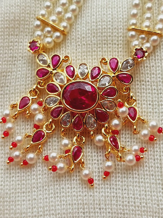 Opulent golden floral pendant with vibrant red and clear gemstones, complemented by strands of lustrous pearls, against a neutral-toned knit fabric background.